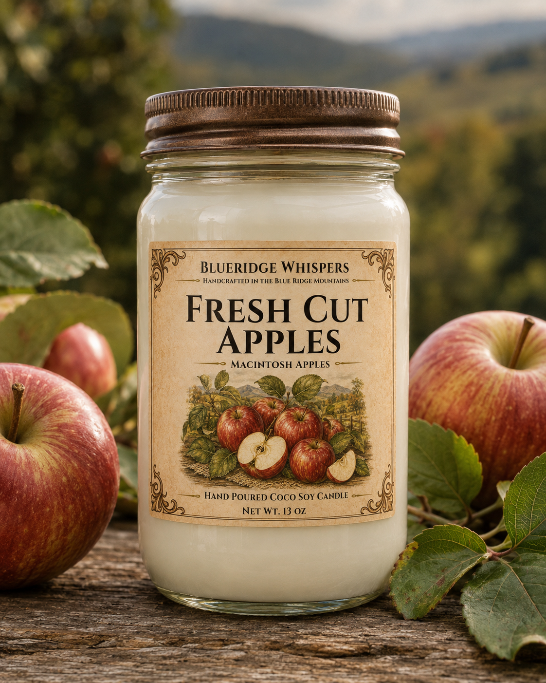 A jar candle labeled 'Fresh Cut Apples' by Blueridge Whispers, set on a rustic wooden surface with red apples and green leaves around it, and a blurred outdoor mountain background.